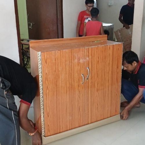 Om Sai packers and movers in Nabinagar safely lifting a large wooden household storage cabinet using protective bottom padding during a residential relocation process.